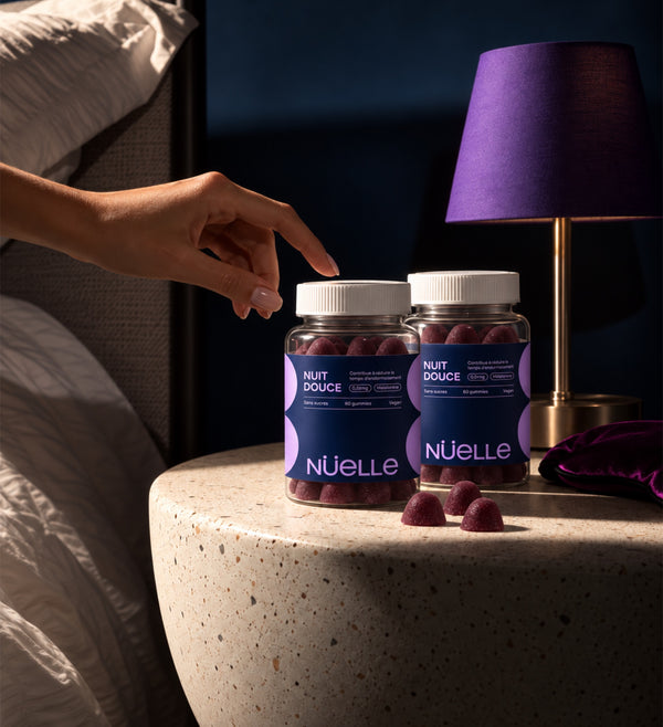 Main qui se tend vers deux pots de gommes NUIT DOUCE posés sur une table de chevet, près d'une lampe violette