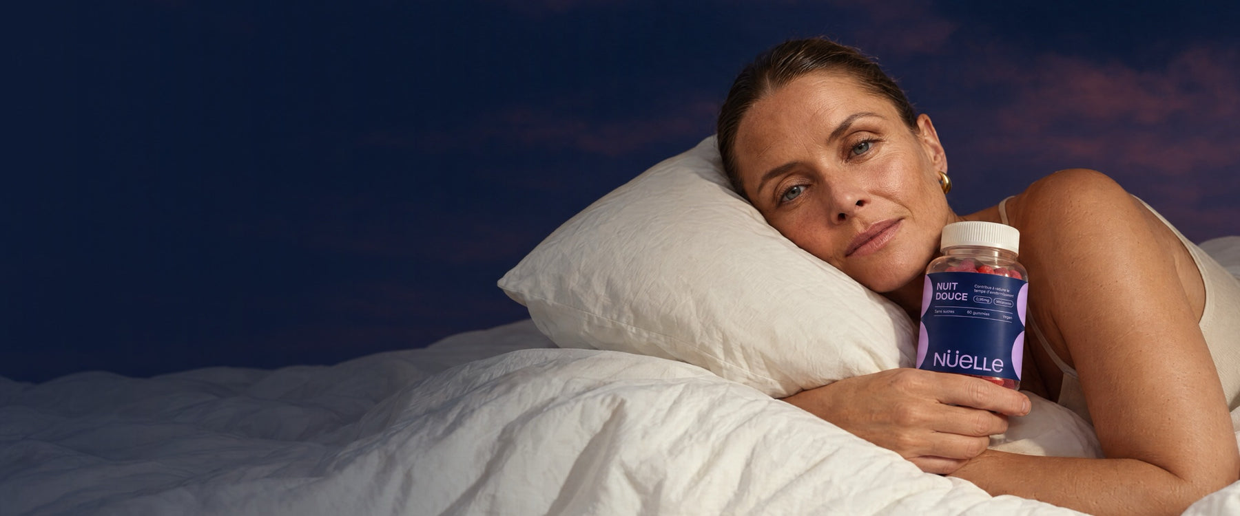 Femme allongée sur un oreiller clair, tenant le pot de gommes NUIT DOUCE de NÜELLE, sous un ciel de nuit